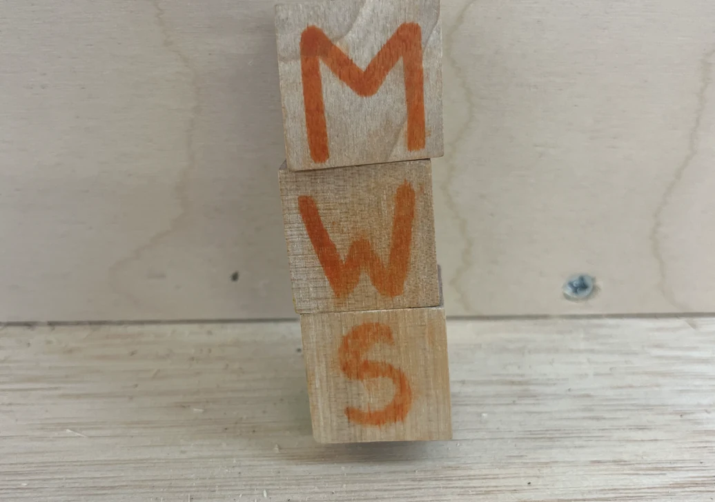 Three decorated wooden pieces standing upright with the letters M, W and S.
