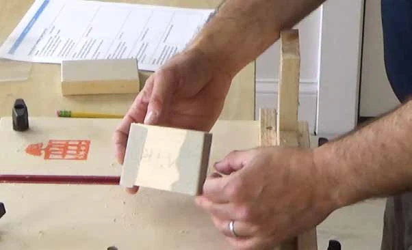 Hands holding a small rectangular wooden sanding block with abrasive paper fitted around its working face.