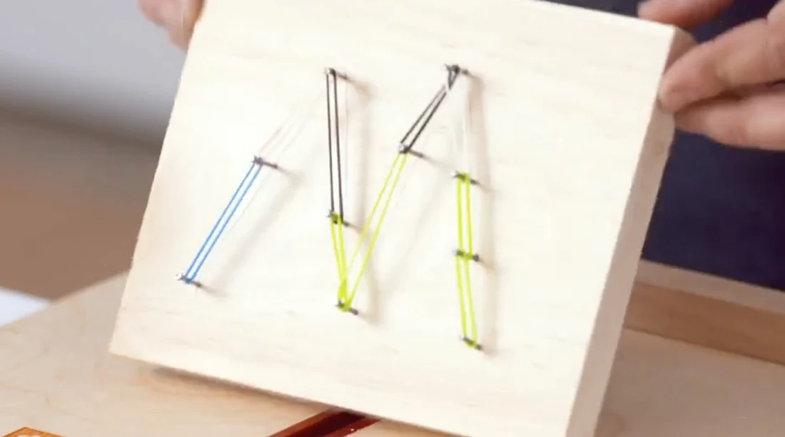 A completed M-shaped pattern made by connecting nails with colorful Rainbow Loom bands on a wooden board.